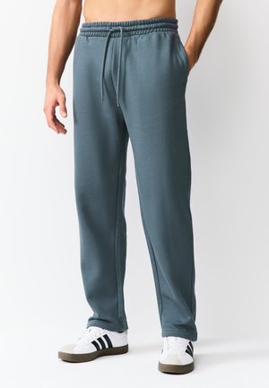 Mand iført skiferblå joggingbukser med elastik i taljen og snøre, kombineret med hvide sneakers med sorte striber.