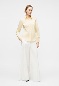 Kvinde står med hænderne i lommerne iført en lysegul langærmet skjorte og hvide bukser med vide ben mod en ensfarvet baggrund.