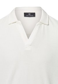 Polo bianco con una trama a costine, con scollo a V e colletto. L'etichetta riporta "Nils Sundström." Design semplice, senza ulteriori motivi.