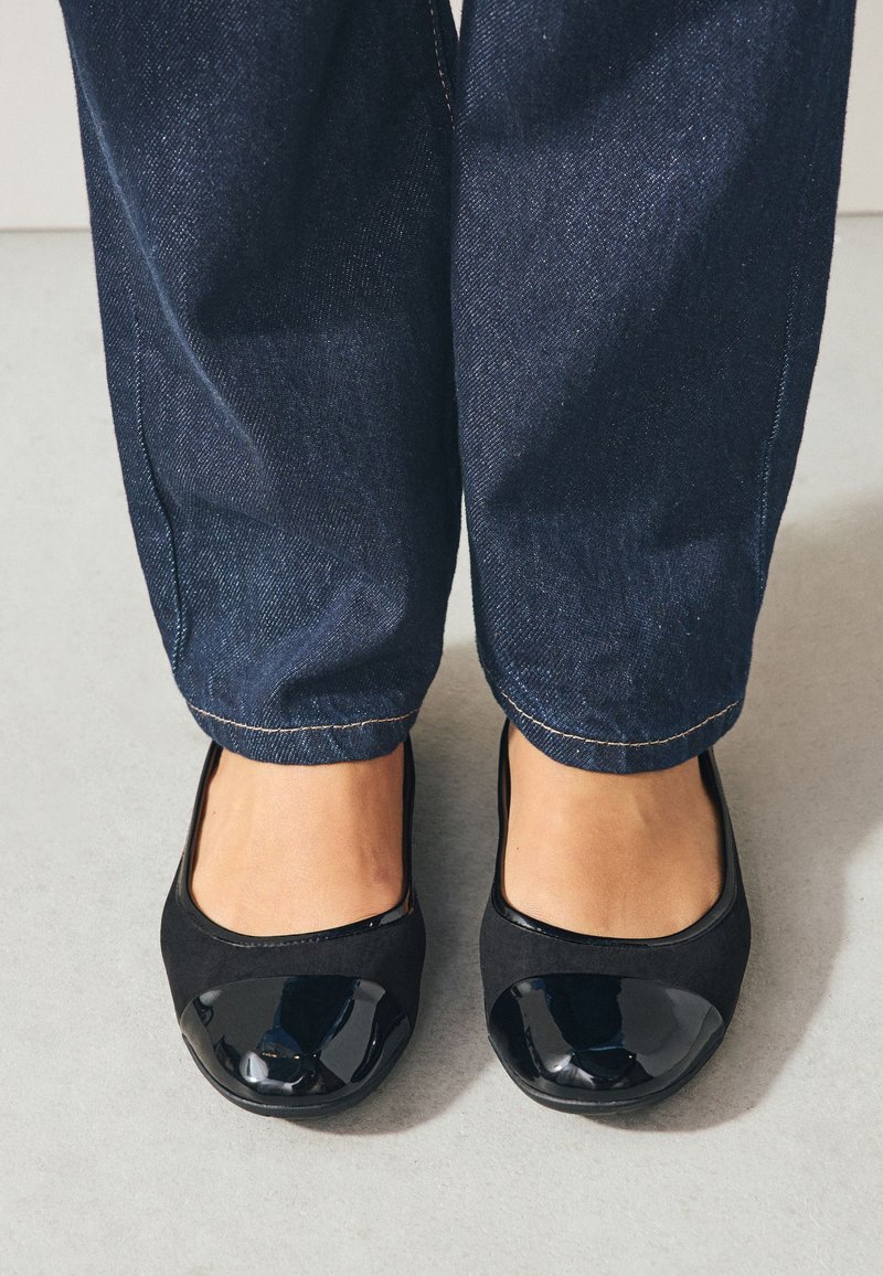 Frau, die dunkelblaue Jeans und schwarze Ballerinas mit glänzenden Zehenkappen trägt, steht auf einem hellgrauen Boden.