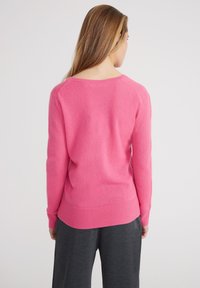 Pull en peluche rose avec un col rond et un ourlet côtelé, présenté sur un modèle vu de dos, associé à un pantalon gris foncé.