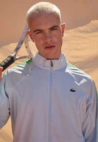 Jachetă sport de culoare albastru deschis cu accente verzi, închidere prin fermoar și textură din plasă. Modelul ține o rachetă de tenis sprijinită de umăr.
