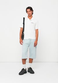 Polo shirt blanc avec logo, short en denim bleu clair et baskets noires. Un sac à bandoulière noir ajoute du détail à la tenue.