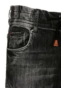 Le jean en denim noir présente une texture lisse, des coutures blanches contrastantes et une poche avant avec une petite étiquette de marque. Une ceinture ajustable est également incluse.