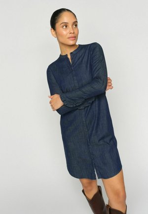 Sieviete ar tumšiem matiem sasietiem augstā bulciņā, valkājot garā piedurkņu džinsa kleitu un brūnus zābakus, stāv ar sakrustotām rokām un skatās uz sāniem.