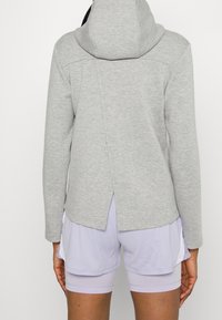 Veste à capuche grise en tissu doux avec un design à dos fendu, associée à un short violet clair, offrant un look athlétique décontracté.