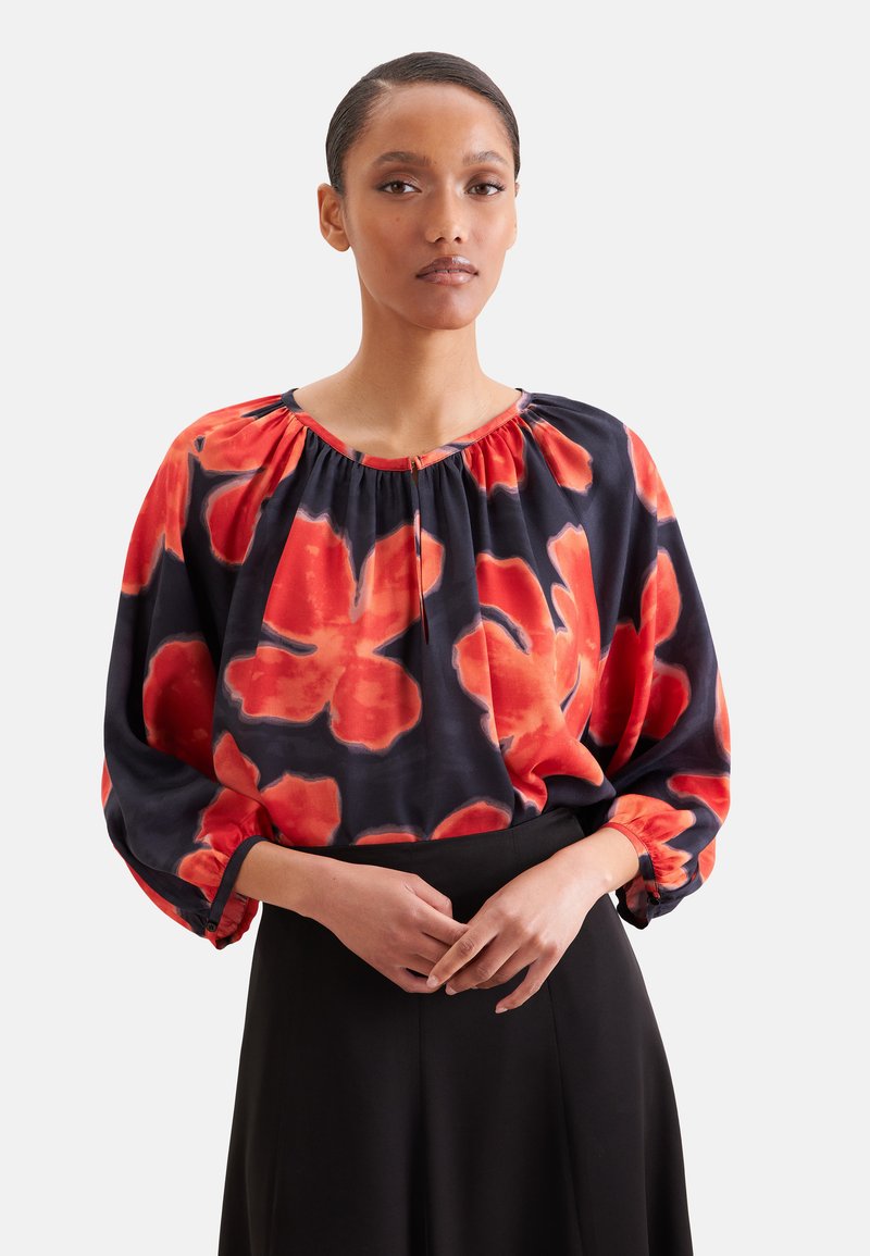 Femme portant un chemisier ample à manches longues avec un grand motif floral rouge et noir et une jupe noire, debout les mains jointes.