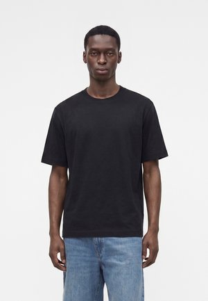 Giovane uomo che indossa una t-shirt nera semplice a maniche corte e jeans azzurri, in piedi su uno sfondo bianco.