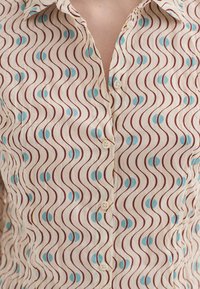 Chemise blanche à boutons avec des lignes ondulées brunes et des formes de croissants bleus répétées portée par une personne.