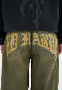 Grüne Jeans mit großen goldenen Buchstaben "ED HARDY" auf der Rückseite, mit einer verblassten Textur und standardmäßigen Taschenplatzierungen.