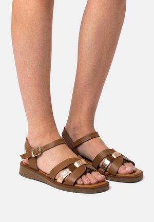 Pieds portant des sandales plates en cuir marron avec des brides à la cheville et des accents métalliques rose doré sur un fond clair.