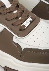 Brązowe i białe sneakersy z teksturowanej skóry, z perforowanym noskiem, brązowymi sznurówkami oraz metalicznym detalem logo na języku. Gładka gumowa podeszwa.