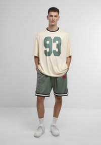 Beige oversized jersey med grønne "93" tryk, parret med grå shorts med sorte striber og et rødt logo-patch. Hvide sneakers på.