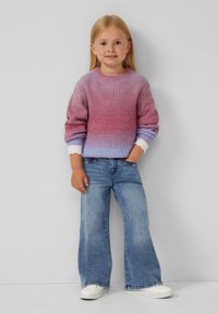 Strickpullover mit einem Farbverlauf von Pink zu Lila, weißen gewellten Bündchen; kombiniert mit weit geschnittenen blauen Jeans und weißen Sneakers.