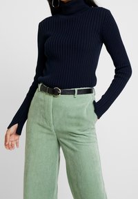 Kobieta w granatowym ribbed golfie z otworami na kciuki oraz w jasnych zielonych spodniach z korduroju z czarnym pasem i złotą klamrą.