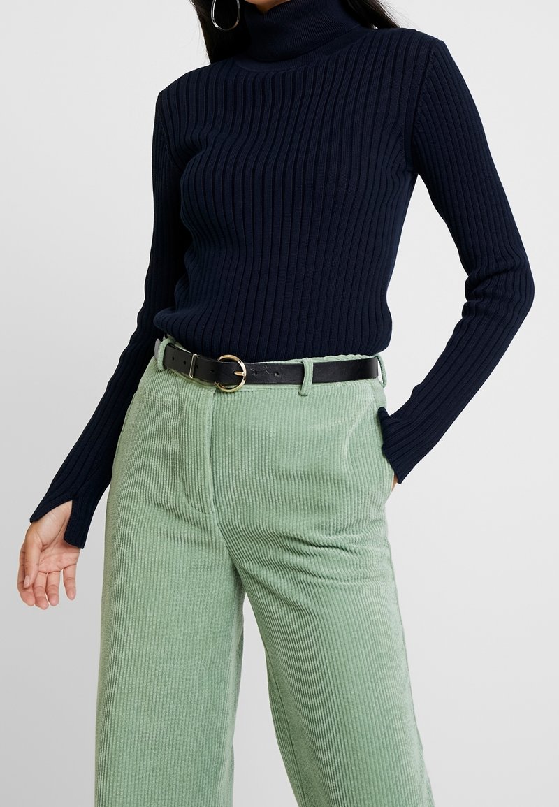 Kobieta w granatowym ribbed golfie z otworami na kciuki oraz w jasnych zielonych spodniach z korduroju z czarnym pasem i złotą klamrą.