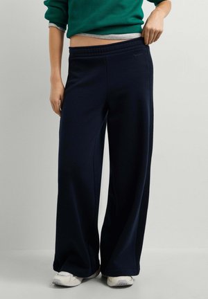 Persoon draagt losse zwarte broek en witte sneakers, houdt tailleband vast om pasvorm te tonen, draagt een groene en grijze gelaagde top.