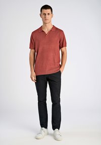 Röd kortärmad polotröja med krage, tillverkad av texturerad stickad tyg, kombinerad med mörka byxor och vita sneakers.