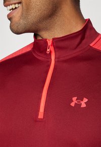 Rødbrun sportslig pullover med høj krave og halvlange lynlås. Har lyserøde accenter på skuldrene og logo på brystet. Glat tekstur.