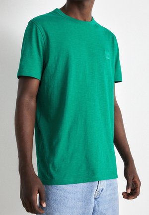 Homme portant un t-shirt vert uni à manches courtes et un jean bleu clair, debout devant un fond blanc uni.