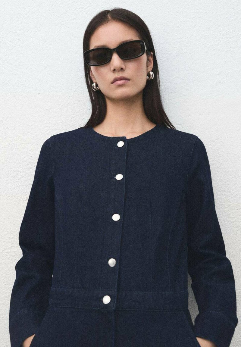 Vestido de mezclilla azul marino con cuello redondo, cinco botones blancos en la parte frontal y mangas largas. Textura suave y diseño ajustado.