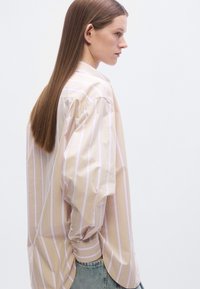 Hellbeiges Hemd zum Knöpfen mit rosa und weißen vertikalen Streifen, lockerer Schnitt, lange Ärmel und abgerundeter Saum. Kombiniert mit blauen Jeans.