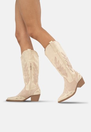 Botas de vaquero de ante beige con recortes en forma de hoja, cremallera lateral y tacón apilado. Presentan una punta afilada y un acabado texturizado.