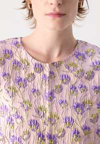 Brodert kjole i lys rosa med intrikate lilla blomster mønstre og gull aksenter. Har rund hals og teksturert stoff.