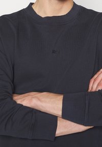 Mörkblå långärmad sweatshirt med ribbat halsringning och ärmslut, med en subtil svart logotyp på framsidan. Mjuk tygtextur.