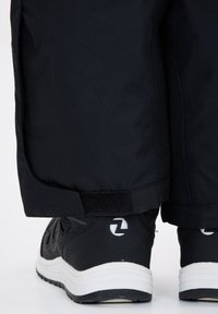 Zwarte waterdichte broek die gedeeltelijk zwarte sport schoenen bedekt, met een gestructureerde bovenkant, witte zool en een logo op de hiel.