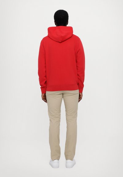 Sudadera roja con una gran capucha ajustable, puños acanalados y dobladillo. Combinada con pantalones beige y zapatillas blancas para un look casual.
