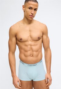 Μυώδης άνδρας που φοράει ανοιχτό μπλε ανδρικά εσώρουχα Calvin Klein, στέκεται μπροστά σε απλό λευκό φόντο, κοιτώντας ελαφρώς προς τα πλάγια.