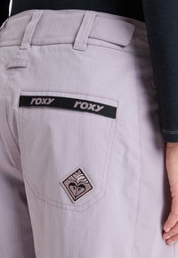 Light purple Roxy pants with a textured pattern, featuring a black waistband, dual back pockets, and an embroidered emblem on the right pocket.