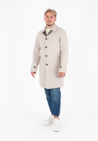Cappotto di lana beige chiaro con colletto a scollo, quattro bottoni frontali e due tasche laterali. Abbinato a jeans blu e sneakers bianche.