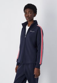 Chaqueta de navidad con cremallera, cuello alto, forro interior blanco y rayas rojas y blancas en las mangas. Incluye un pequeño logotipo en el pecho.