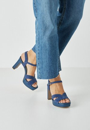 Blue suede high-heeled sandals with crossed straps and ankle buckle. Square toe and block heel design. Worn with light blue denim pants.