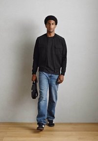 Jachetă bomber neagră, tricou negru, blugi de culoare albastru deschis și adidași negri. Ținând o geantă neagră, stând pe o podea din lemn cu un perete gri în spate.