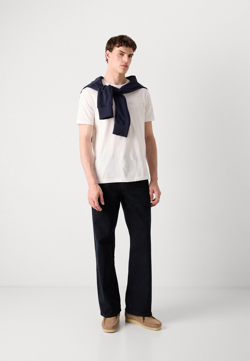 Weißes Baumwoll-T-Shirt mit gesticktem Logo, kombiniert mit dunkelblauen lässigen Hosen und einem marineblauen Sweatshirt, das über die Schultern gelegt ist. Braune Schuhe.