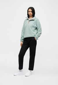 Γυναίκα που στέκεται με το ένα χέρι στην τσέπη, φοράει ανοιχτό μπλε φλις πουλόβερ, μαύρο παντελόνι, λευκά αθλητικά παπούτσια και λευκές κάλτσες μπροστά σε μονόχρωμο φόντο.