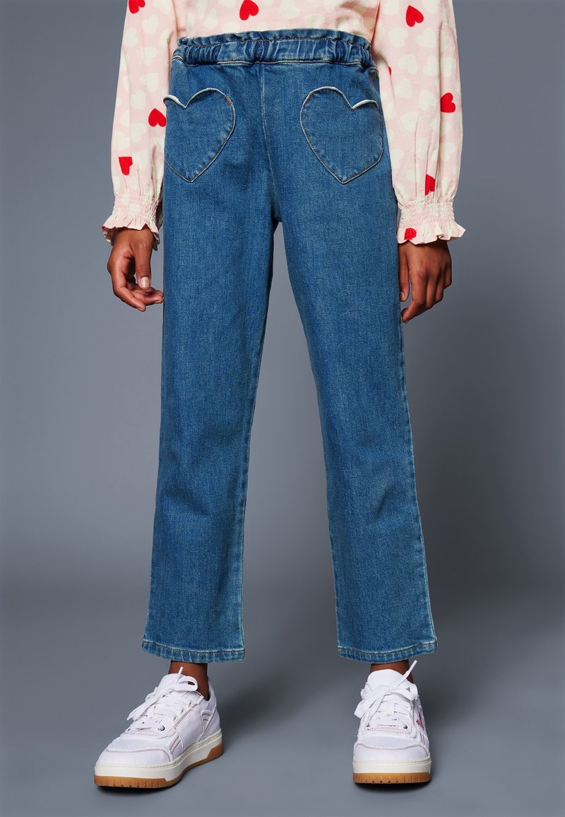 Enfant portant un jean bleu avec des poches arrière en forme de cœur, des baskets blanches et un t-shirt à manches longues rose avec des motifs de cœurs rouges.