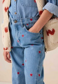 Jean en denim avec broderie de cœur rouge, caractérisé par une taille haute et une coupe décontractée, associé à un gilet en polaire beige avec des accents rouges.