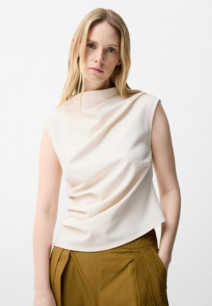 Femme aux longs cheveux blonds portant un haut crème sans manches avec un tissu drapé et un pantalon plissé couleur moutarde, debout sur fond blanc.