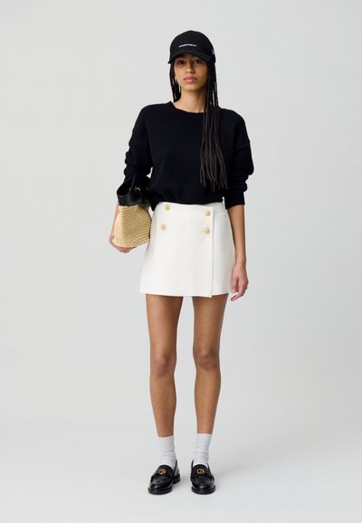 Suéter negro, falda corta blanca de doble botonadura, mocasines negros, calcetines grises, gorra negra, sosteniendo una bolsa de paja, de pie contra un fondo claro.