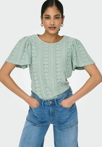 Hellgrünes Oberteil mit Puffärmeln, Augenmuster und horizontal gestreiftem Design, kombiniert mit hochgeschnittenen blauen Jeans.