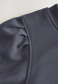 Grijze gebreide pofmouwdetail op een geribde manchet en schouderpaneel, die de stoffenstructuur en stiksels van dichtbij toont.