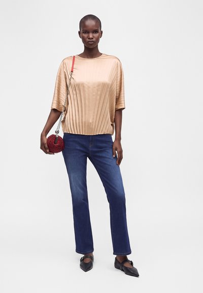Femme portant un top en satin beige plissé, un jean bleu, des chaussures noires pointues, et tenant un petit sac rouge perlé avec une bandoulière chaîne.