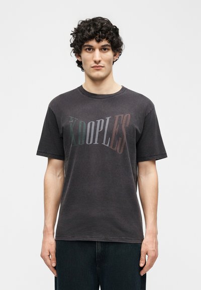 Νεαρός με κατσαρά μαλλιά που φοράει μια ξεθωριασμένη μαύρη μπλούζα με το λογότυπο "THE KOOPLES", στέκεται μπροστά σε ένα απλό λευκό φόντο.