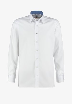 Homme portant une chemise blanche boutonnée rentrée dans un pantalon bleu marine, tenant une veste noire sur l'épaule, debout devant un fond uni.