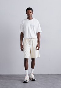 Hvit oversized t-skjorte med en liten blå tekstdetalj; kremfargede shorts med snor; lyse joggesko med detaljer; hvite sportssokker.