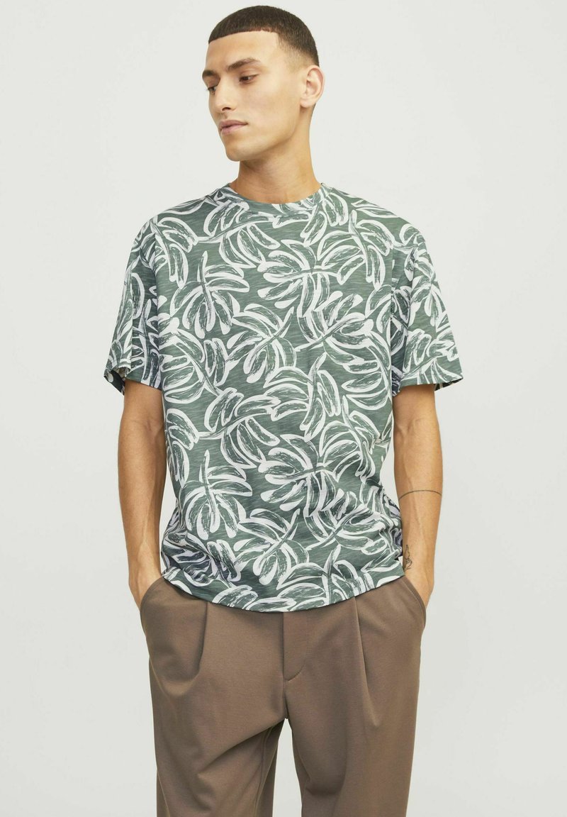 Groene T-shirt met korte mouwen, voorzien van een wit bladerenpatroon op een gestructureerde stof. Model draagt een bruine broek en heeft de handen in de zakken.